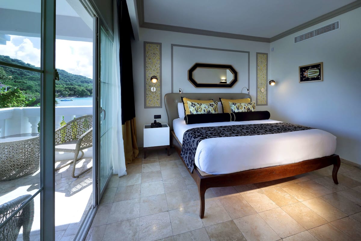 A bedroom with a large bed, a nightstand, a wall-mounted mirror, and a view of a balcony with a chair.