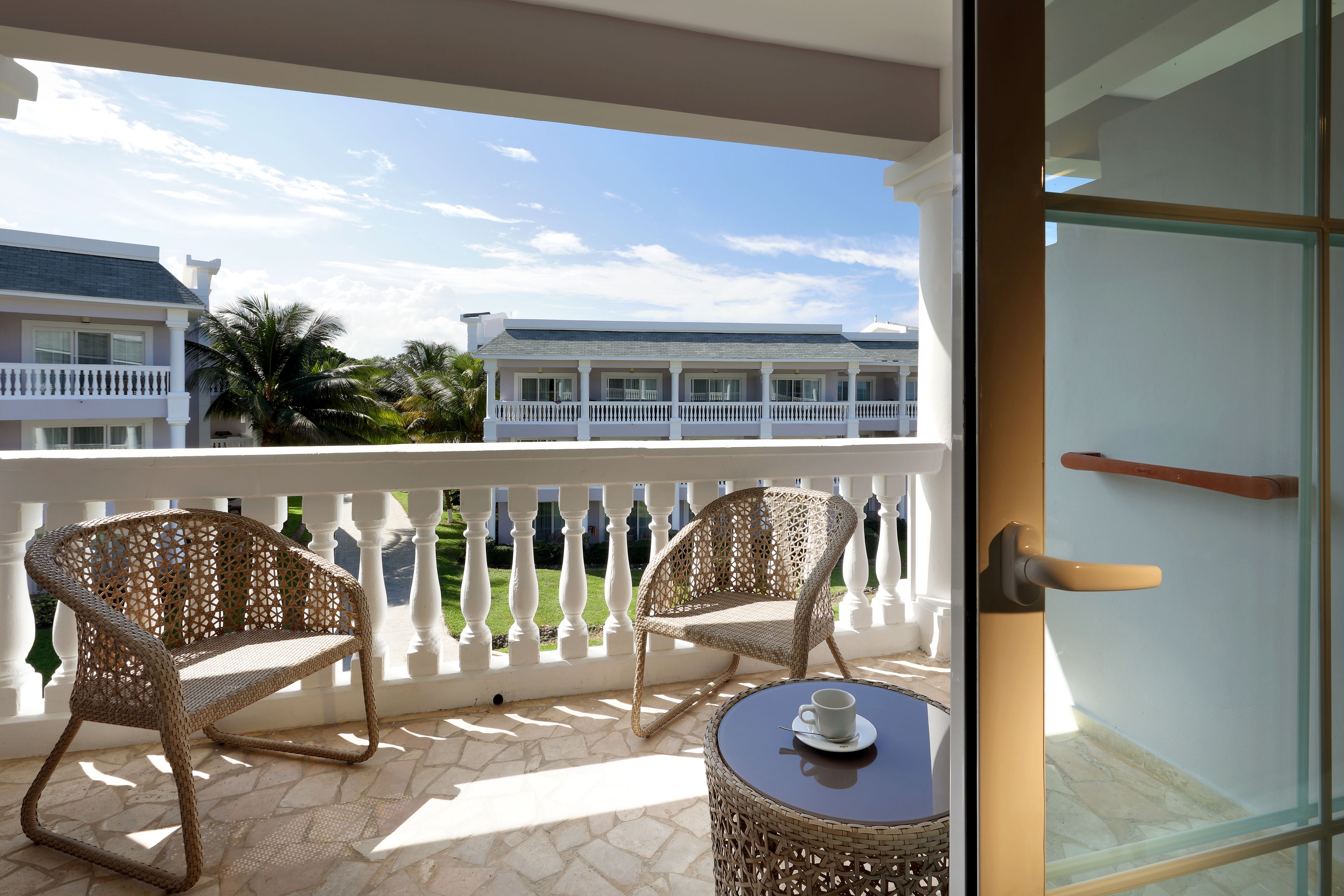 A balcony with wicker chairs and a table, overlooking a pool and buildings.