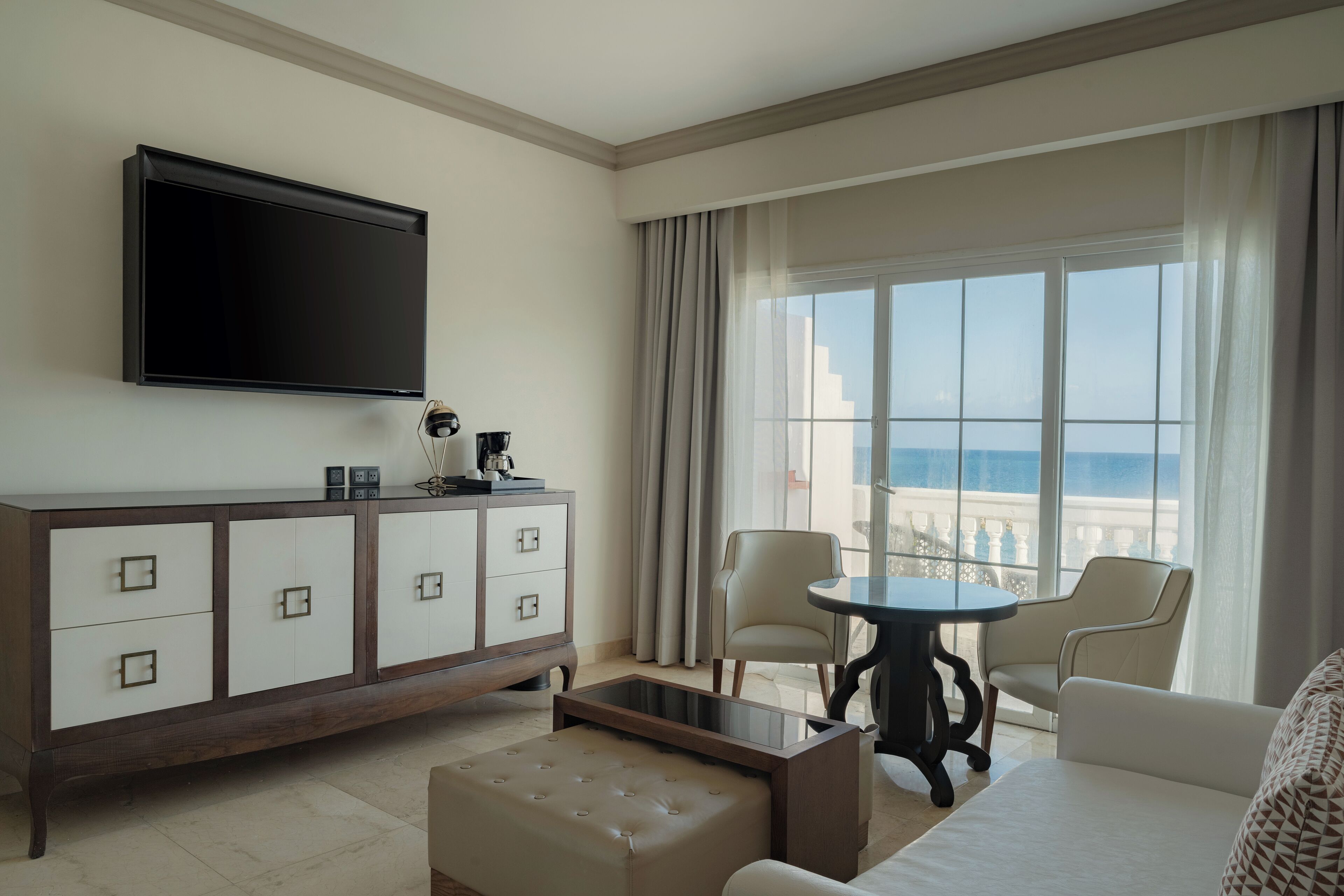 A hotel room with a flat-screen TV, a minibar, a sofa, a coffee table, and a view of the ocean.