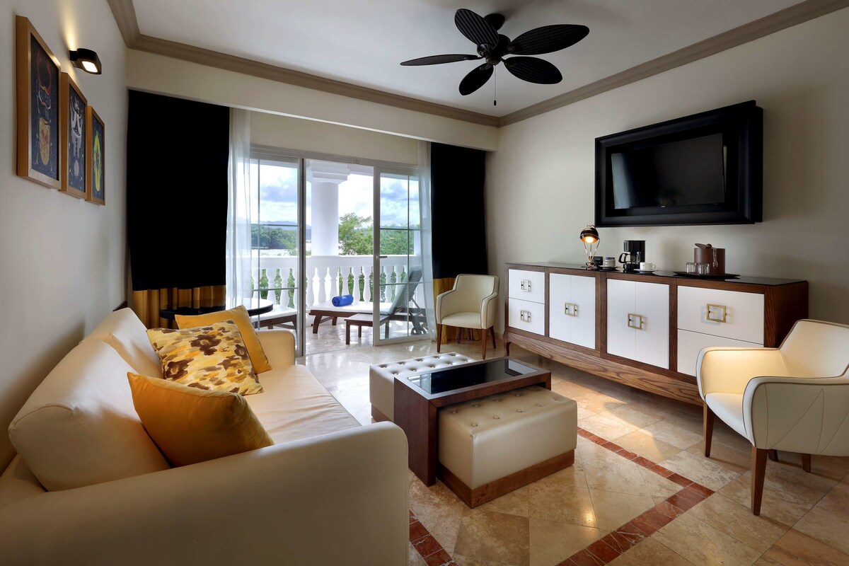 A modern living room with a sofa, ottoman, and a coffee table. There is a television mounted on the wall and a ceiling fan.
