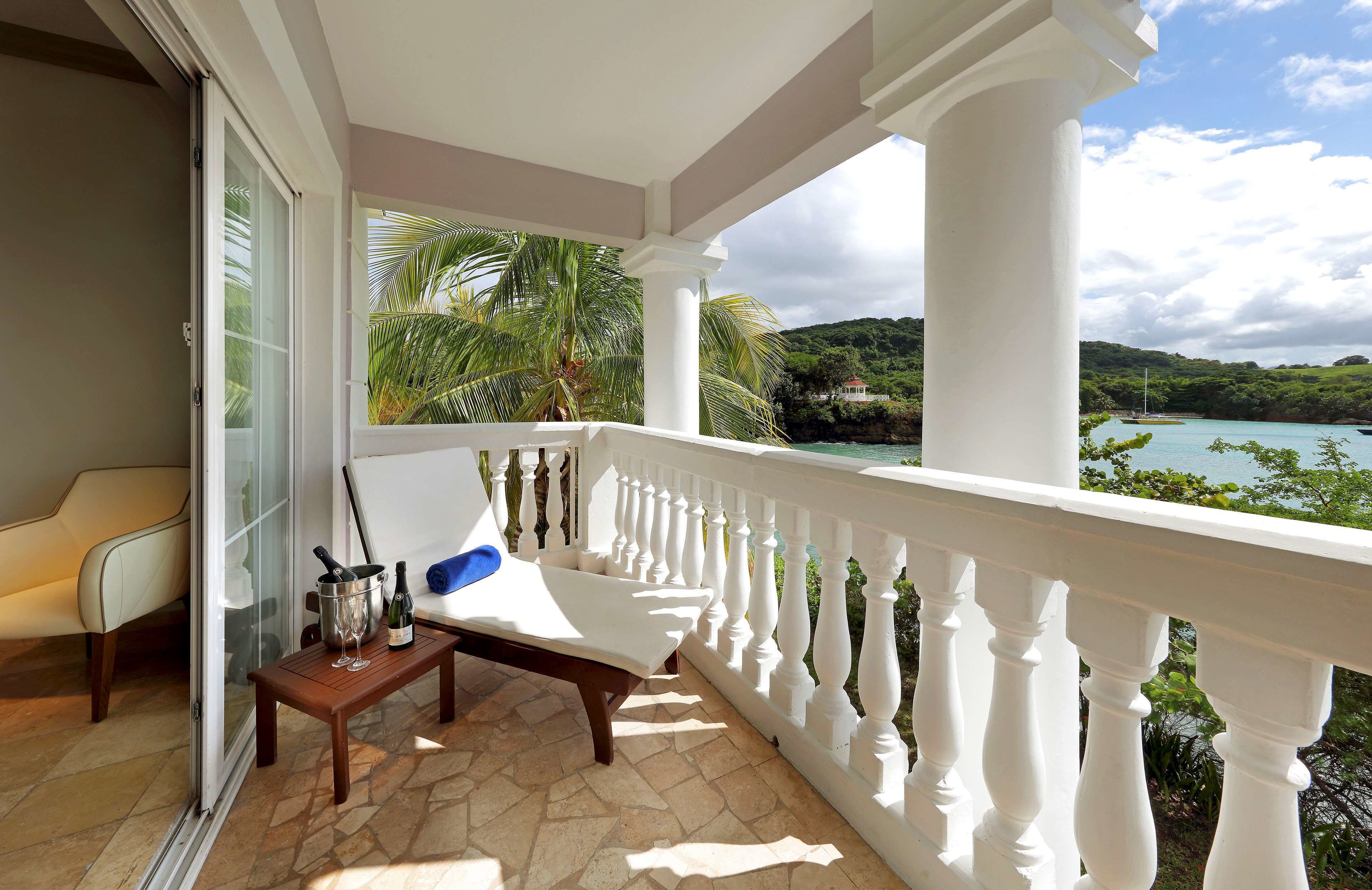 A balcony with a white balustrade, a cushioned chair, a small table with a bottle and glasses, and a view of a coastal landscape with greenery and a body of water.