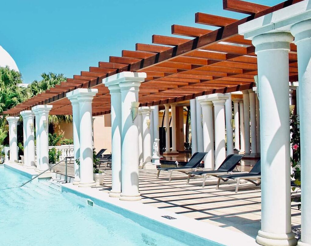 A poolside area with a pergola, white columns, and lounge chairs.