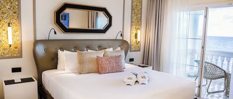 A hotel room with a large bed, a mirror, a nightstand, and a view of the sea through a balcony door.