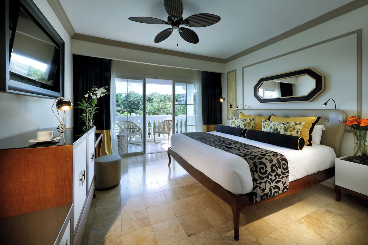A hotel room with a bed, a desk, a chair, a TV, a ceiling fan, and a balcony with a view of trees.