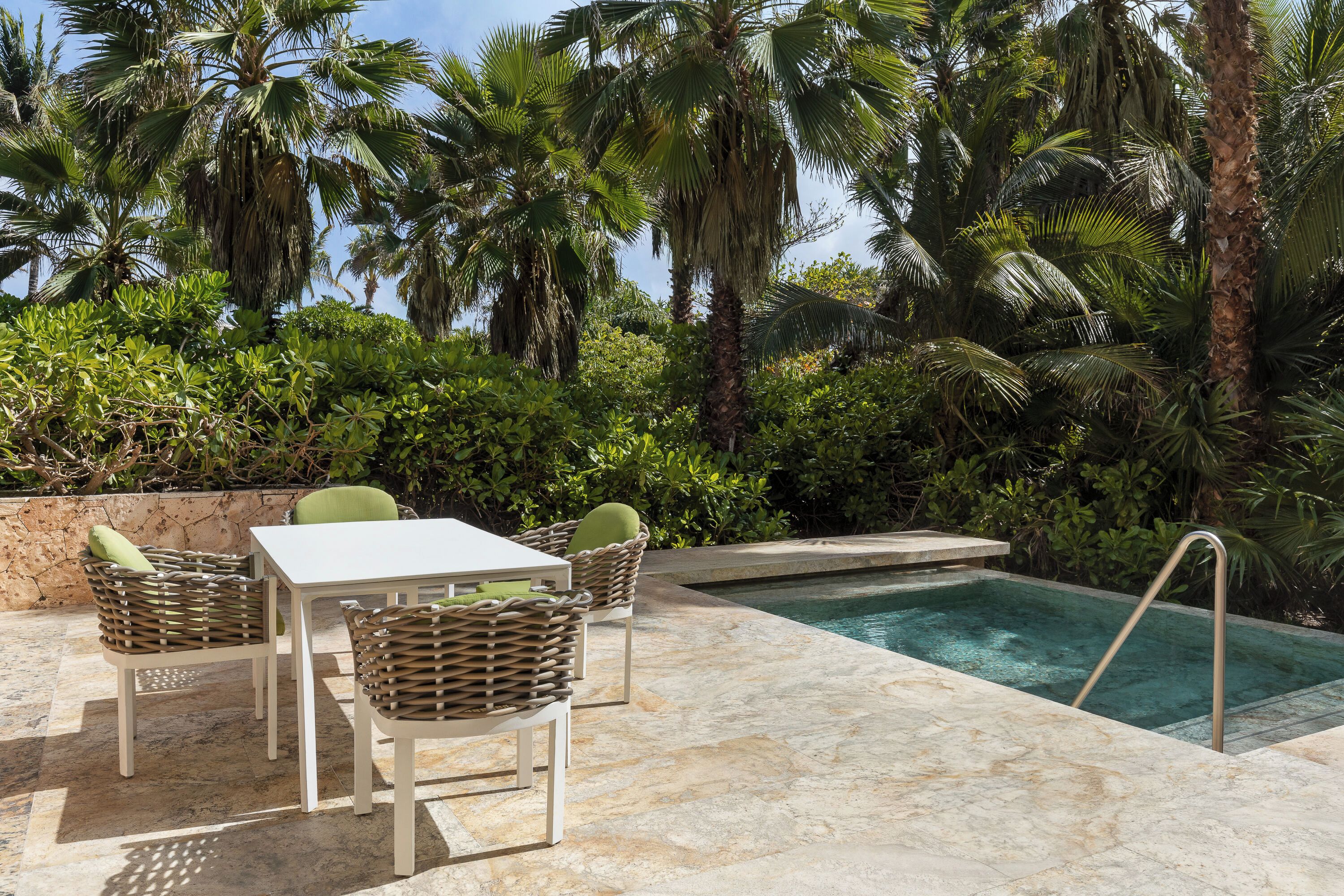 An outdoor pool area with a table and chairs, surrounded by lush greenery and palm trees.