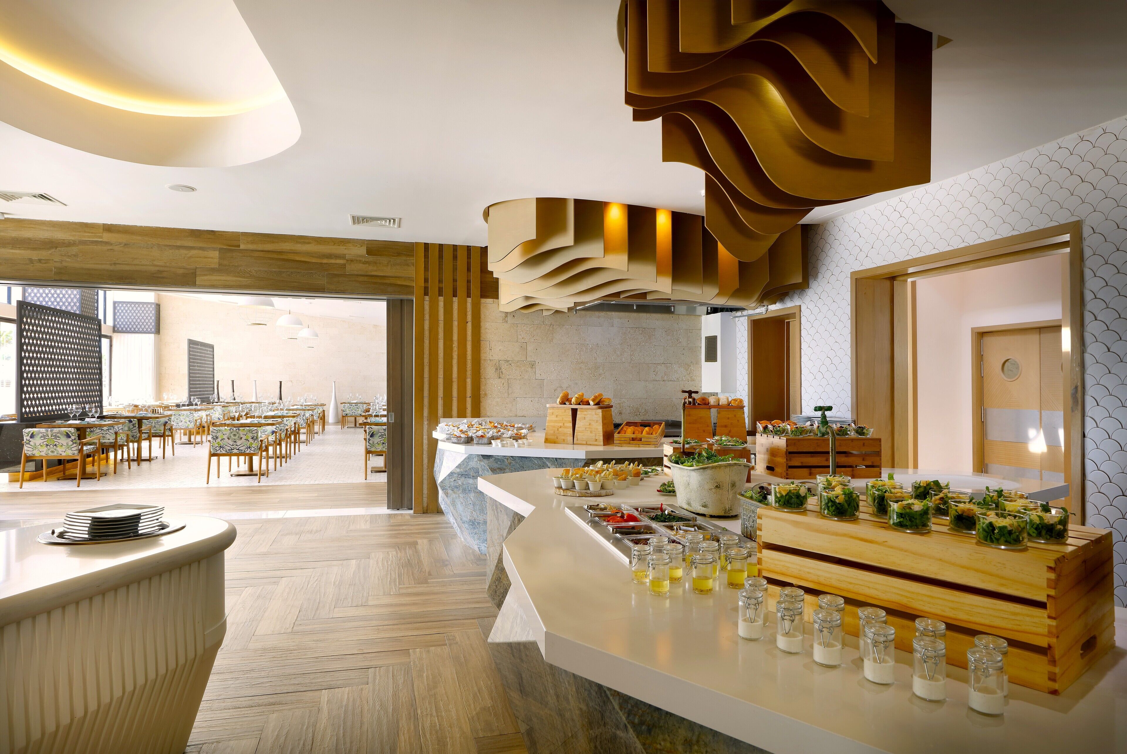 A spacious dining area with multiple tables and chairs, a buffet counter with various food items, and a modern ceiling design.