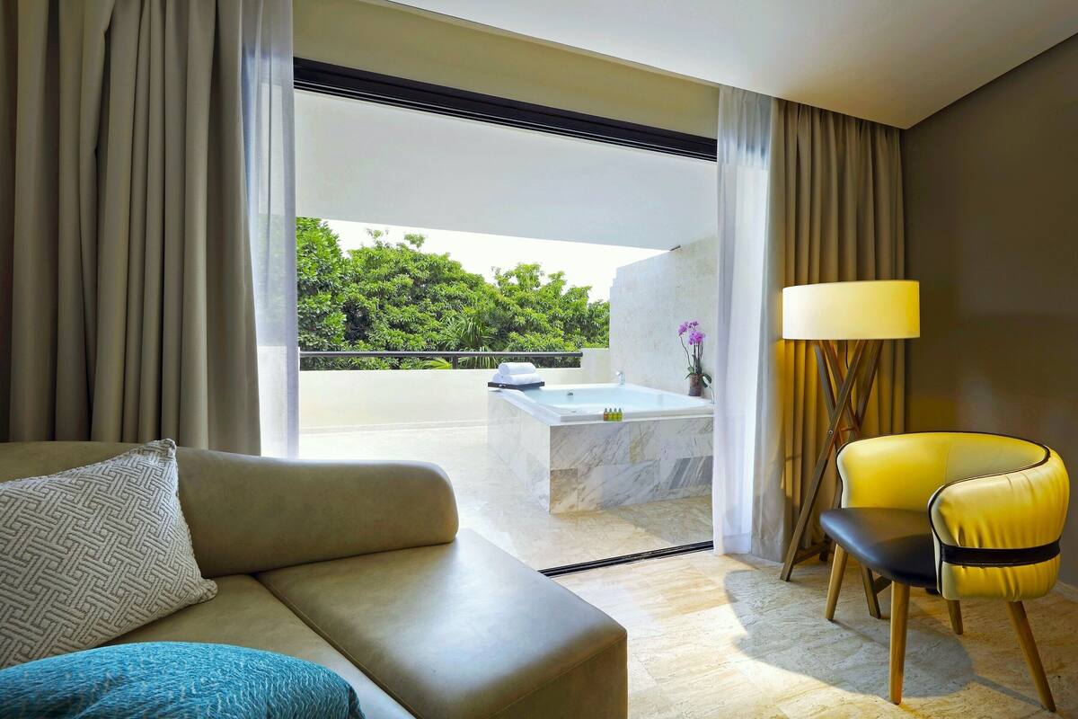 Habitació d'hotel moderna amb sofà, cadira, làmpada i vistes a un balcó amb banyera i vegetació.