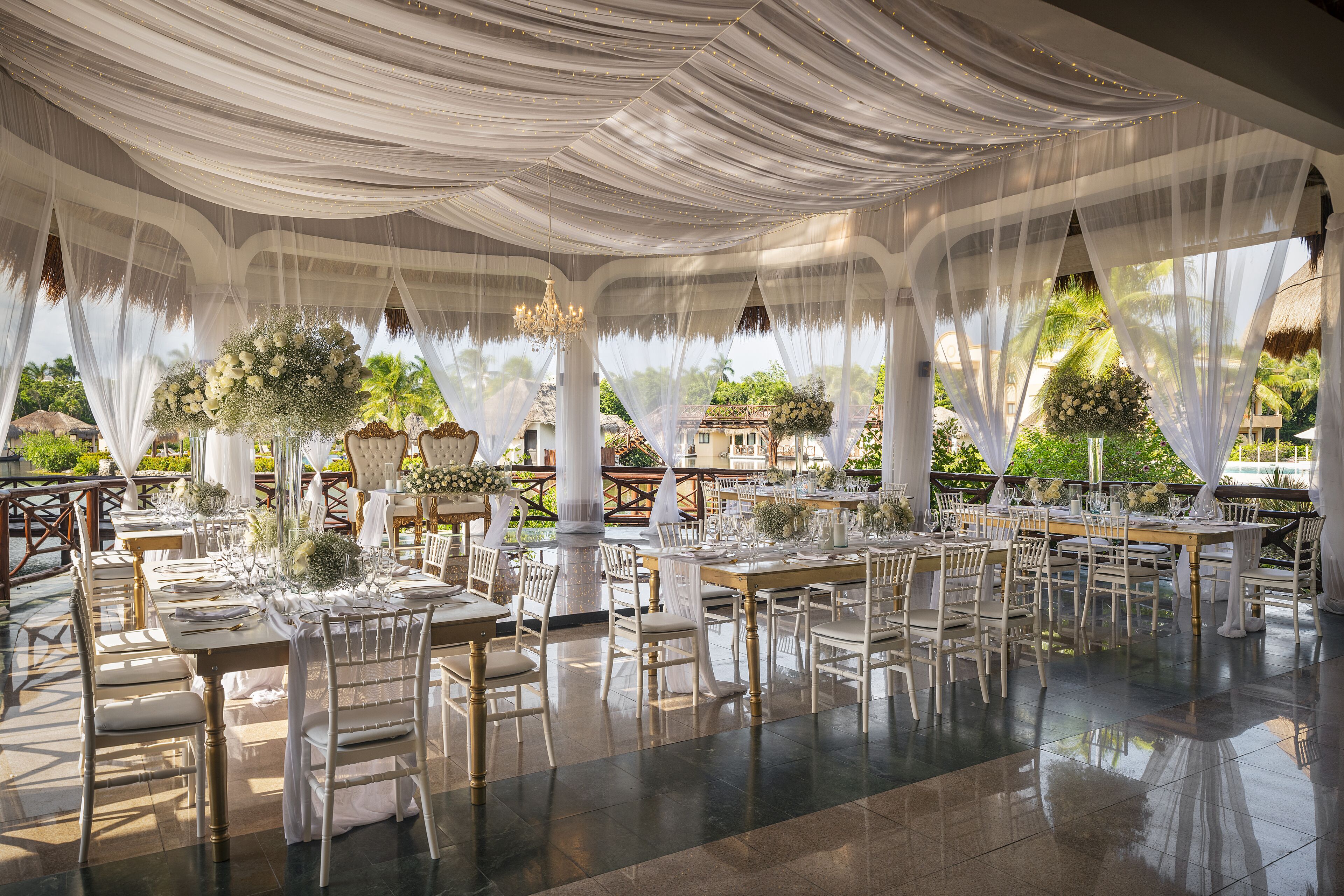 An outdoor dining area with white tables and chairs, draped with white fabric, and surrounded by lush greenery.
