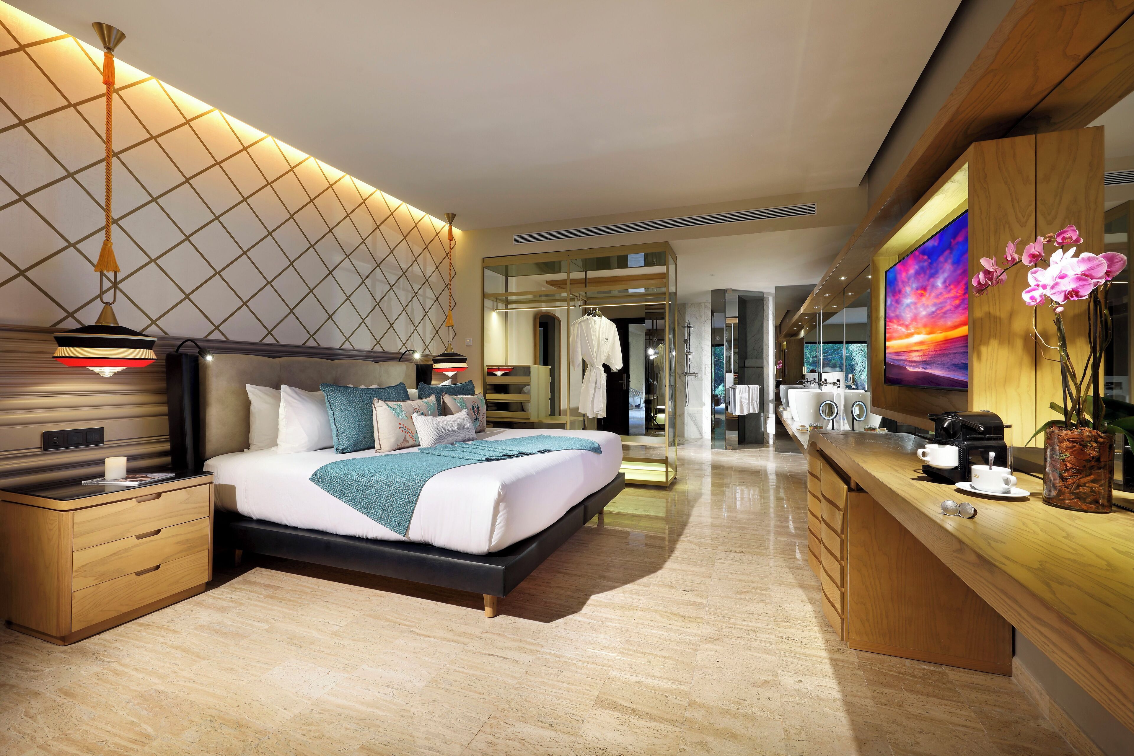A modern hotel room with a large bed, bedside tables, a vanity with a mirror, and a television displaying a sunset.