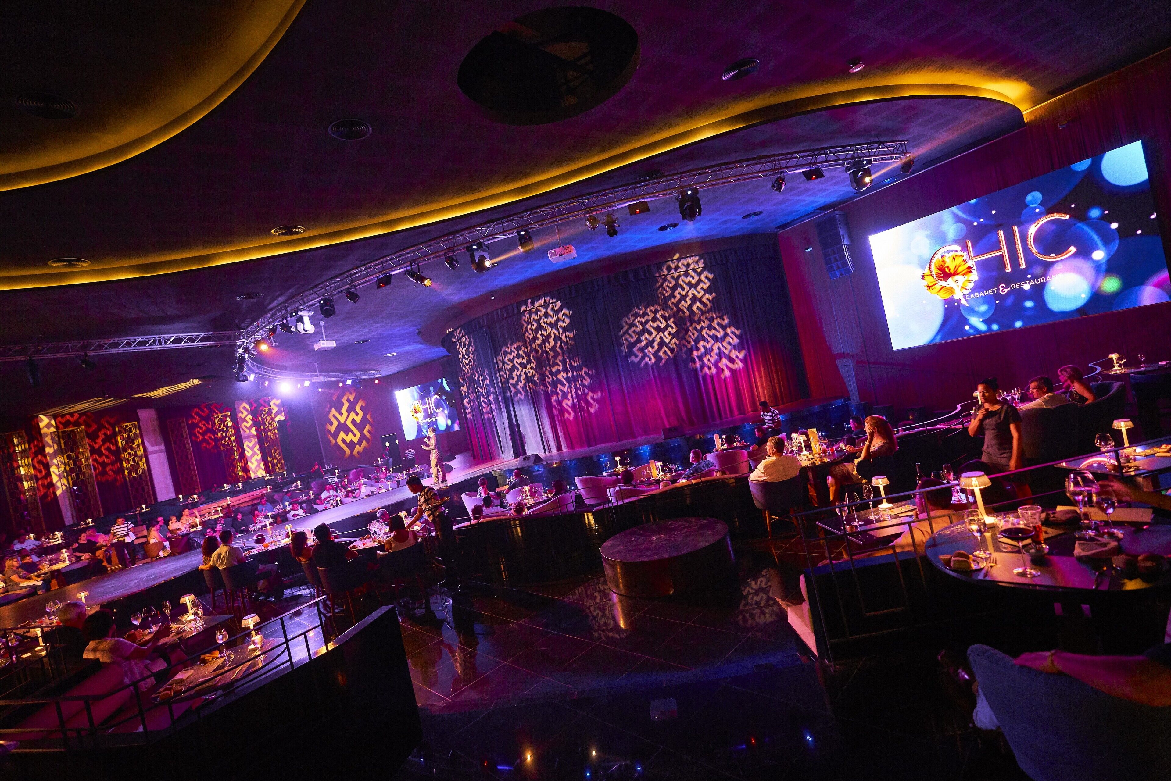 A spacious indoor venue with a stage, seating areas, and a large screen displaying the logo "HIC".