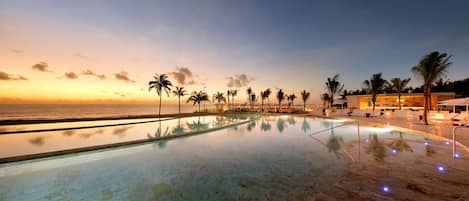 A serene infinity pool with a clear view of the ocean at sunset, surrounded by palm trees and lounge chairs.