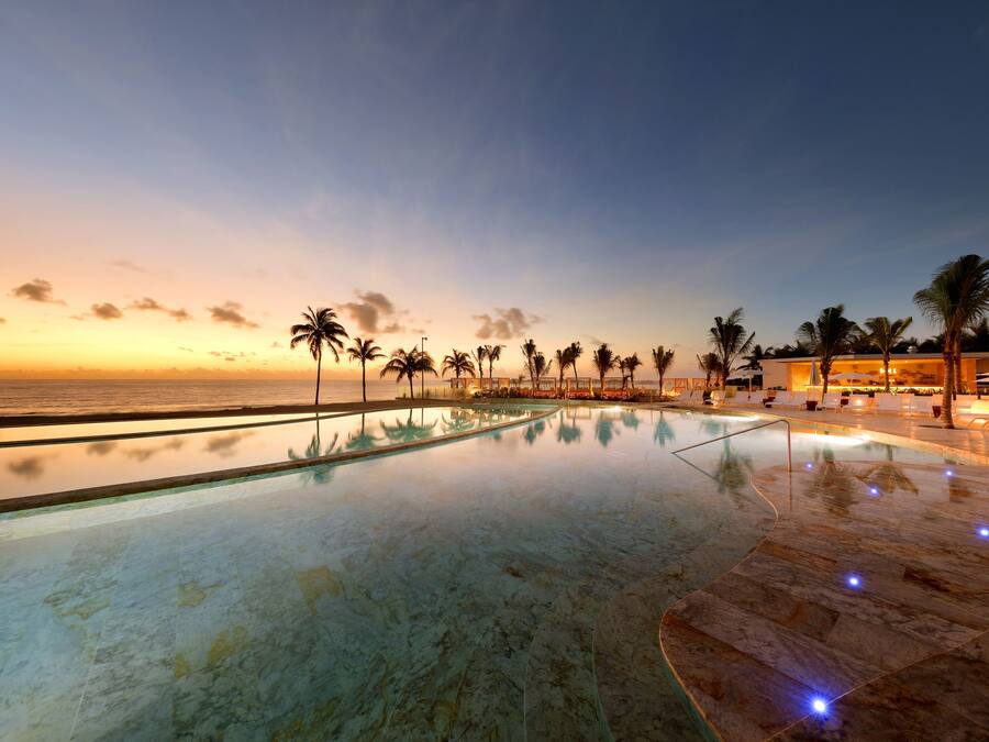 Ein ruhiger Infinity-Pool mit freiem Blick auf den Ozean bei Sonnenuntergang, umgeben von Palmen und Liegestühlen.