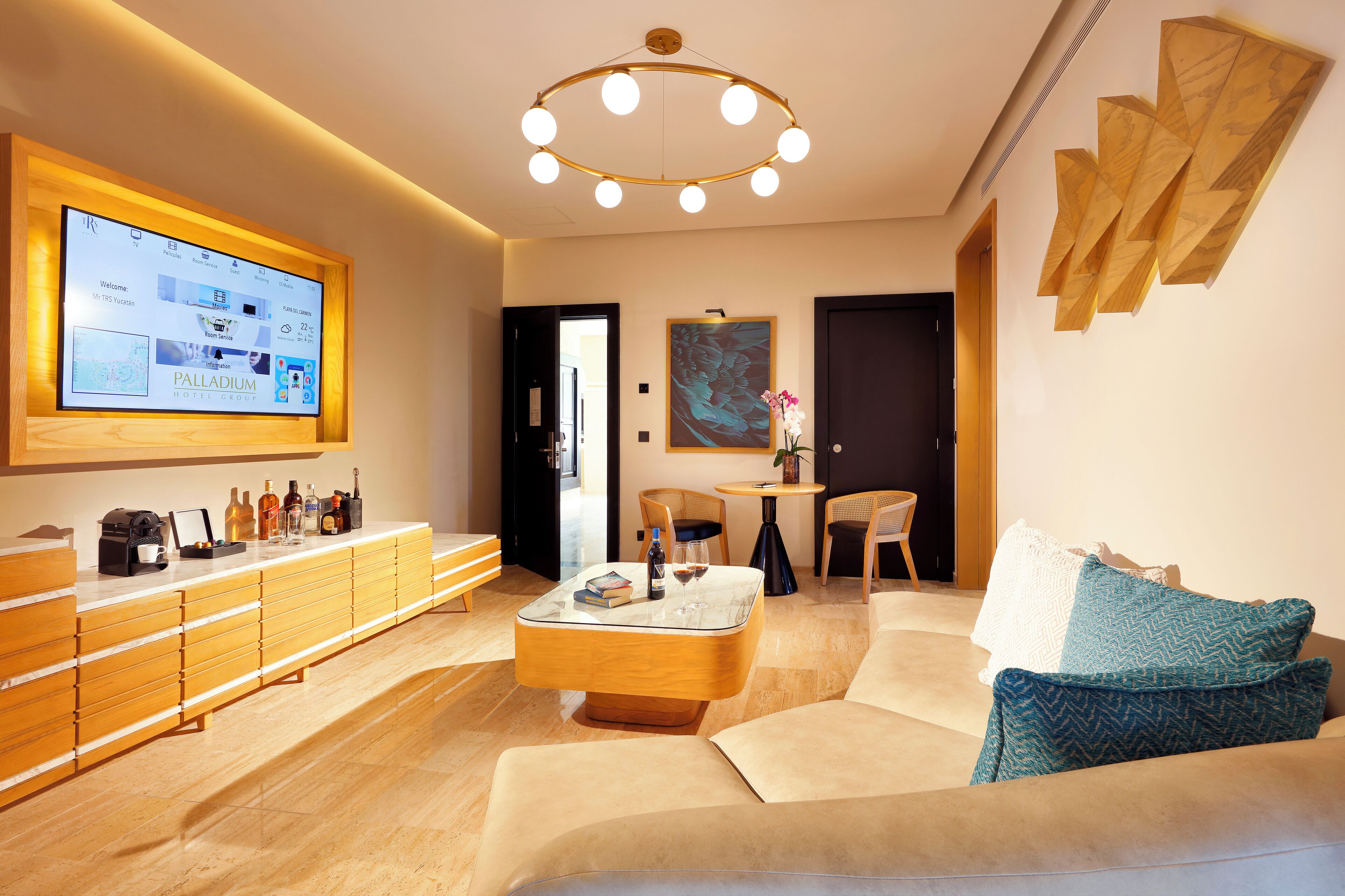 A modern hotel lobby with a large digital display, a wooden reception desk, a round coffee table, and a seating area with a sofa and chairs.