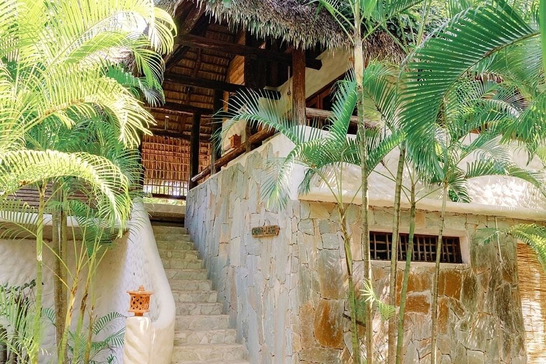 Un escalier en pierre mène à une structure en bois surmontée d’un toit de chaume, le tout étant entouré de palmiers.