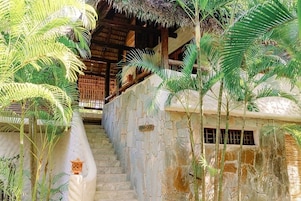 A stone staircase leading to a wooden structure with a thatched roof, surrounded by palm trees.