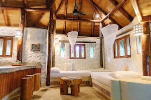 Un interior rústico de madera con una cama, dos taburetes de madera, una mesita y un ventilador de techo.
