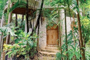 Un edificio de piedra con puerta de madera, rodeado de abundante vegetación y estructuras de bambú.