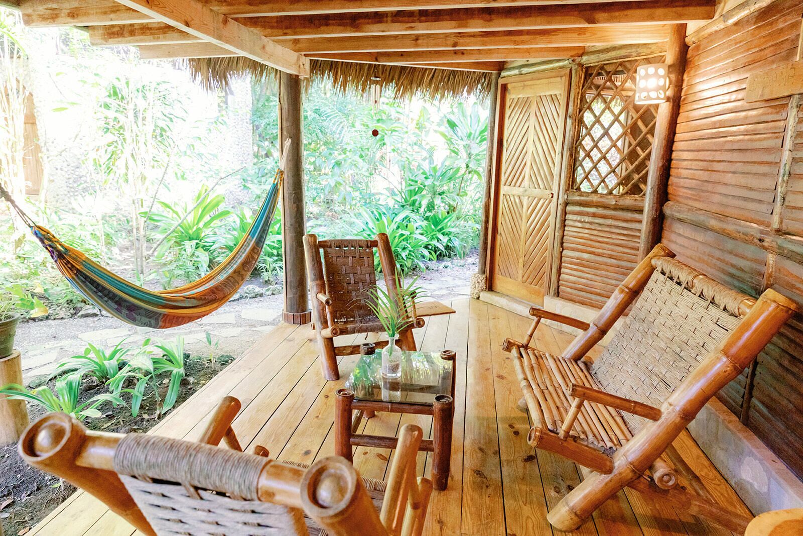A wooden deck with a hammock, two woven chairs, a small table, and a bamboo chair.