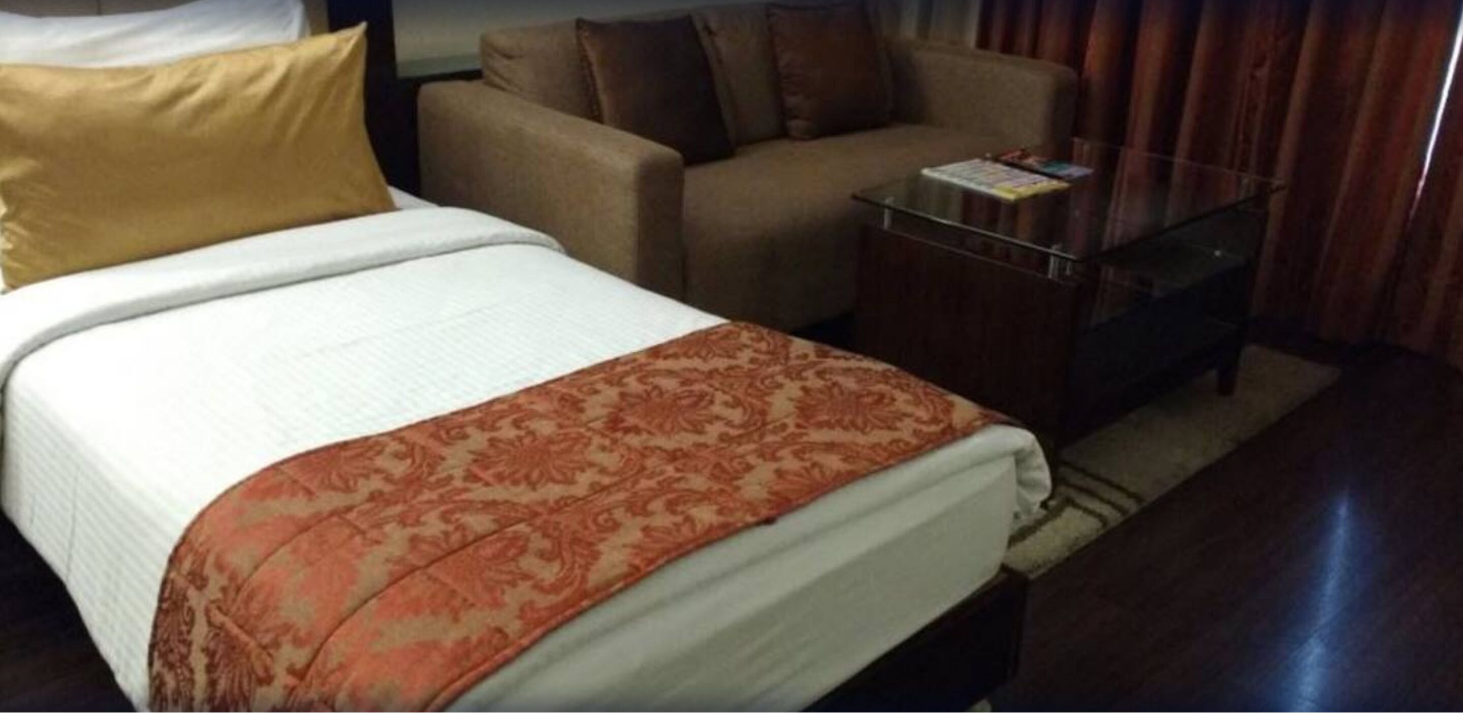 A hotel room with a bed, a sofa, a glass coffee table, and a small table with a magazine.