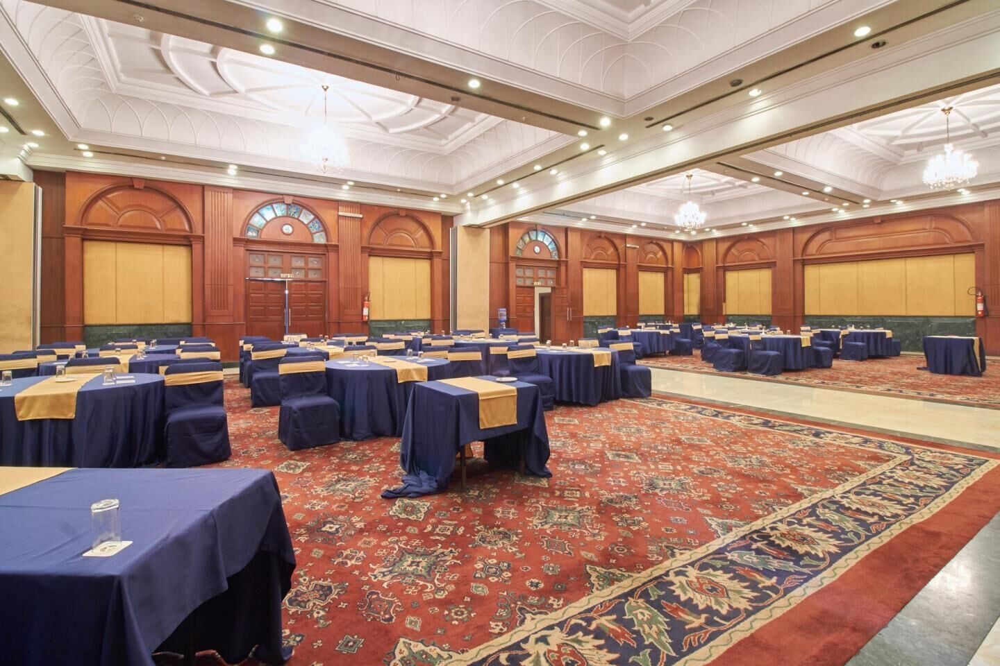 Un amplio salón de conferencias con filas de mesas y sillas, una alfombra estampada y un techo alto con candelabros.