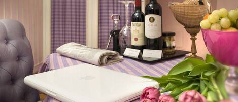 Una computadora portátil sobre una mesa a rayas con una botella de vino, una copa, un bol de uvas y flores rosadas.