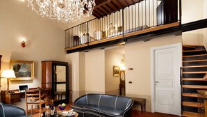 A spacious living room with a black leather sofa, a wooden coffee table, and a chandelier. A staircase leads to an upper level with a balcony. - Graziella Patio Hotel (Arezzo)