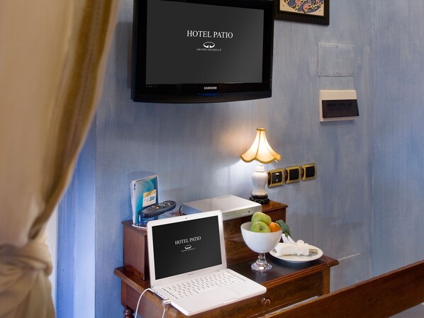 A hotel room with a laptop on a wooden desk, a television mounted on the wall, a bedside table with a lamp, and a fruit bowl.