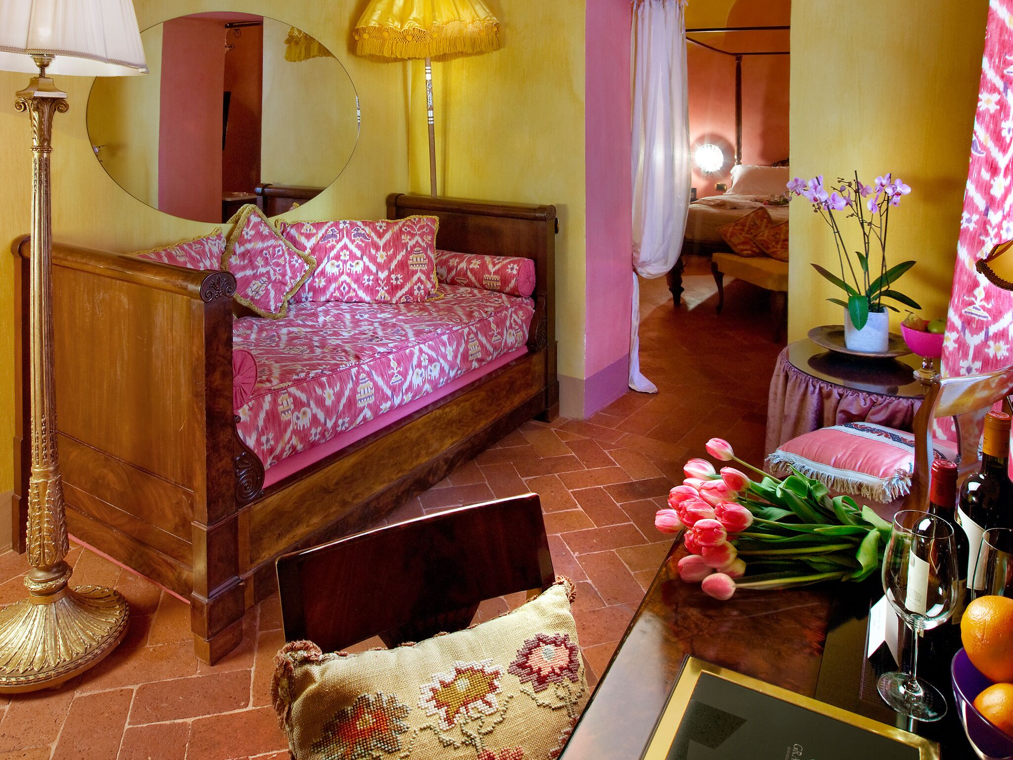 A cozy bedroom with a wooden bed, a mirror, a floor lamp, and a small table with a vase of flowers.