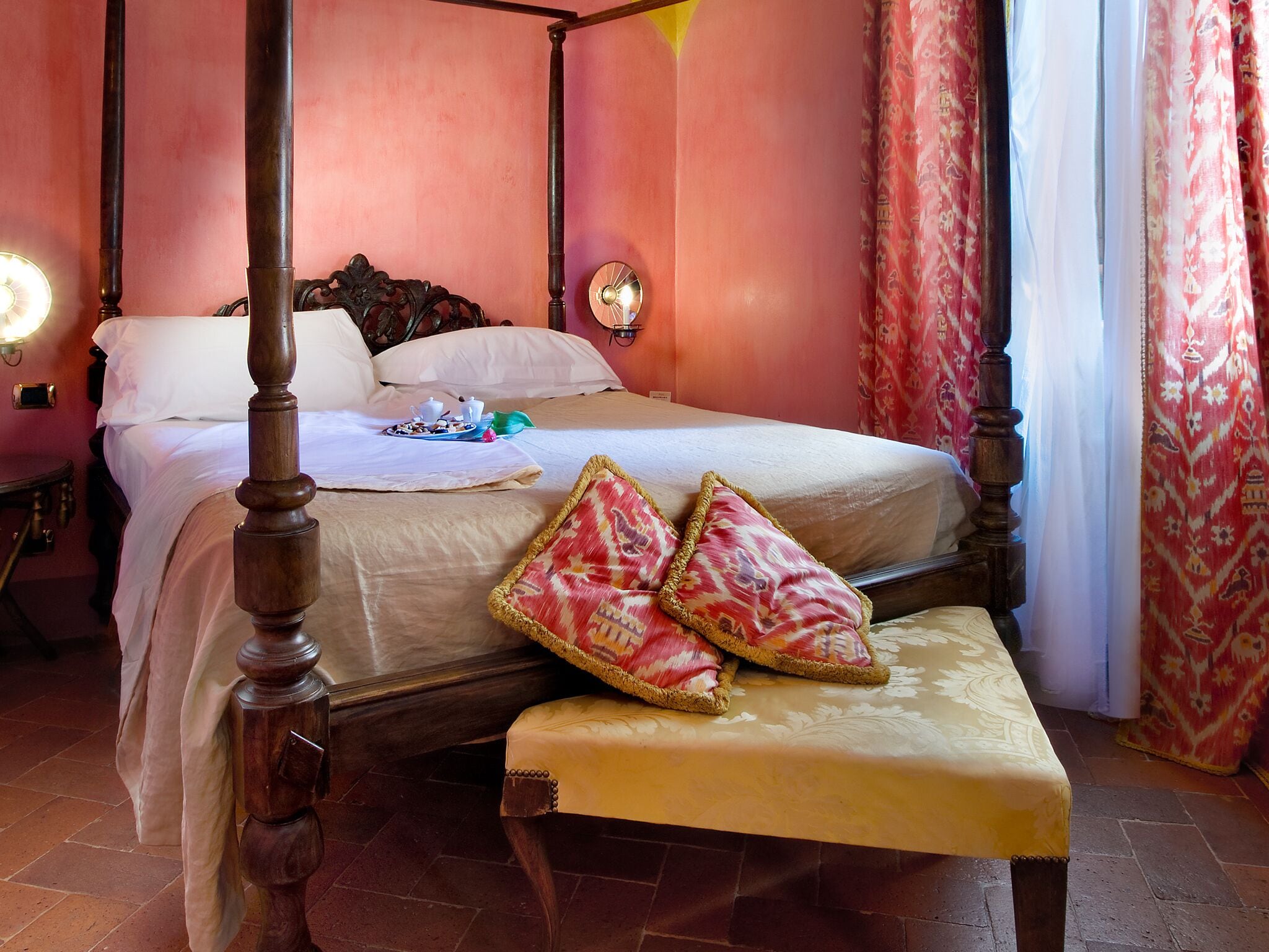 A four-poster bed with a bench, a bedside table, and a wall-mounted lamp.