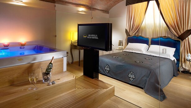 A hotel room with a wooden ceiling, a large bed with a blue headboard, a TV, a jacuzzi, and a bottle of champagne. - Graziella Patio Hotel (Arezzo)