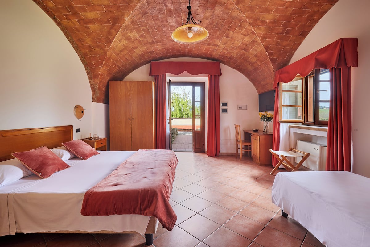 A bedroom with a large bed, a smaller bed, a desk, and a chair. There is a window with curtains, a door leading outside, and a brick ceiling.