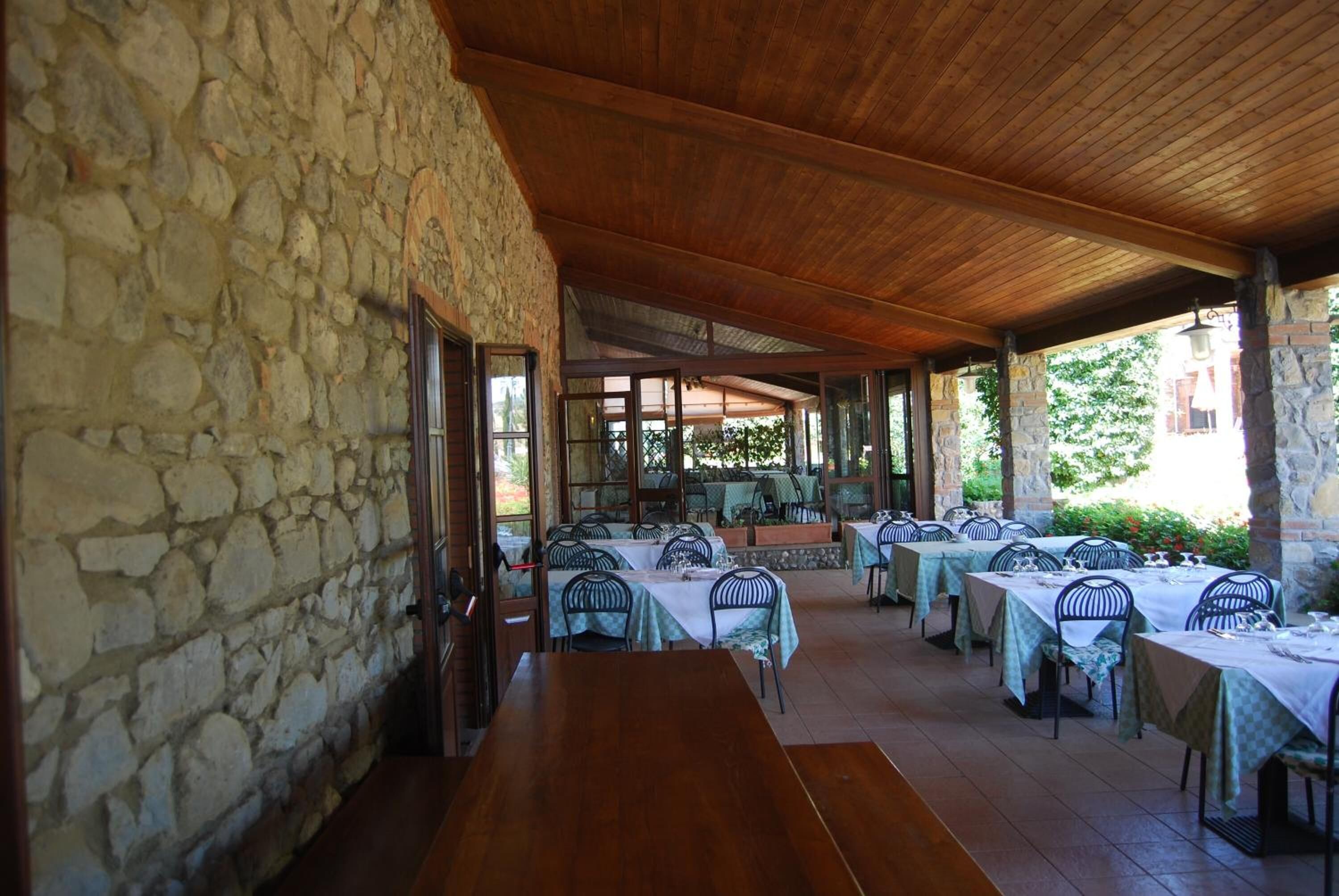 A covered outdoor dining area with stone walls and wooden beams, featuring multiple tables and chairs.