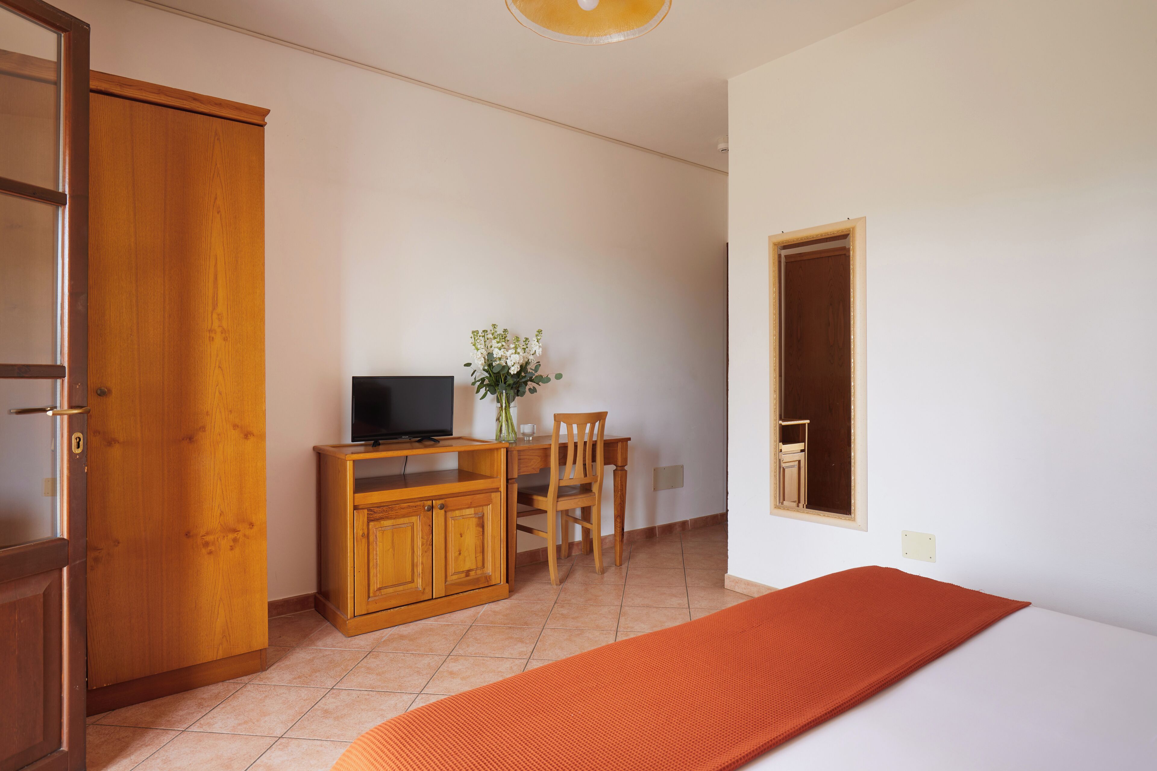 Een slaapkamer met een bed, een houten bureau met een televisie, een stoel en een tafeltje met een bloevas.