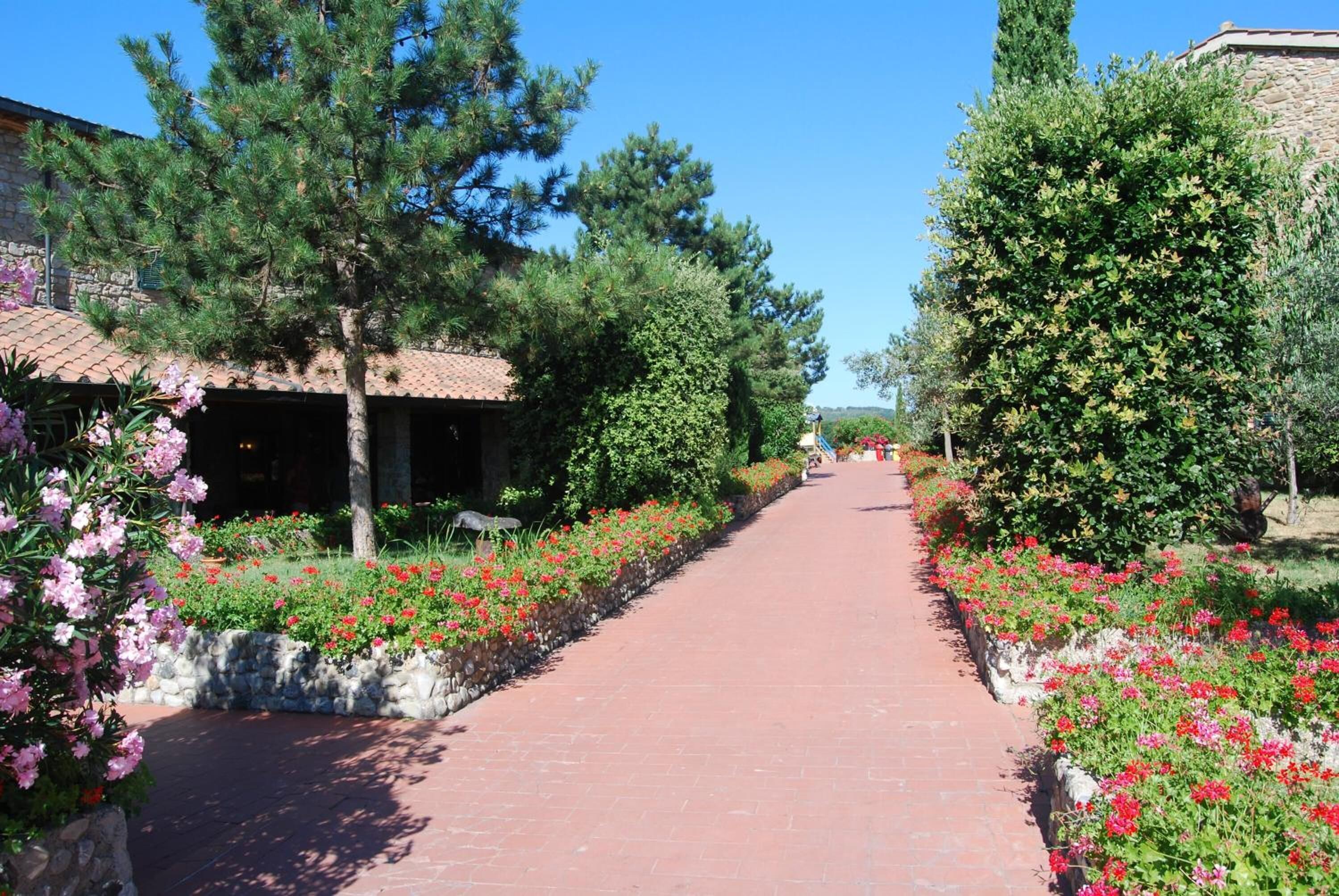 A paved pathway flanked by vibrant flower beds and trees, leading towards a building with a tiled roof.