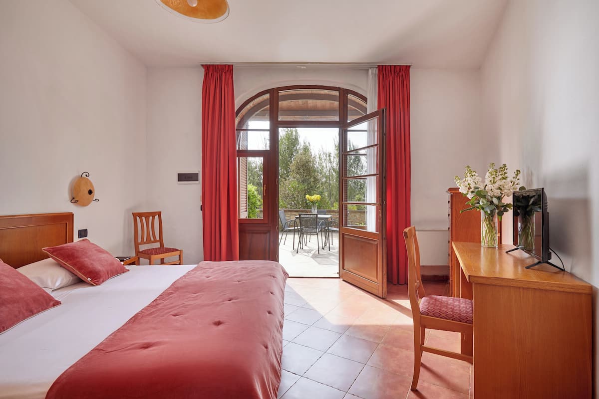 A hotel room with a large bed, a desk, a chair, and a view of the outdoors through a glass door.