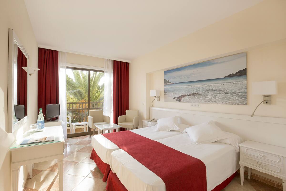 Une chambre d’hôtel avec un lit, un bureau, une chaise, une télévision et un tableau représentant une plage.