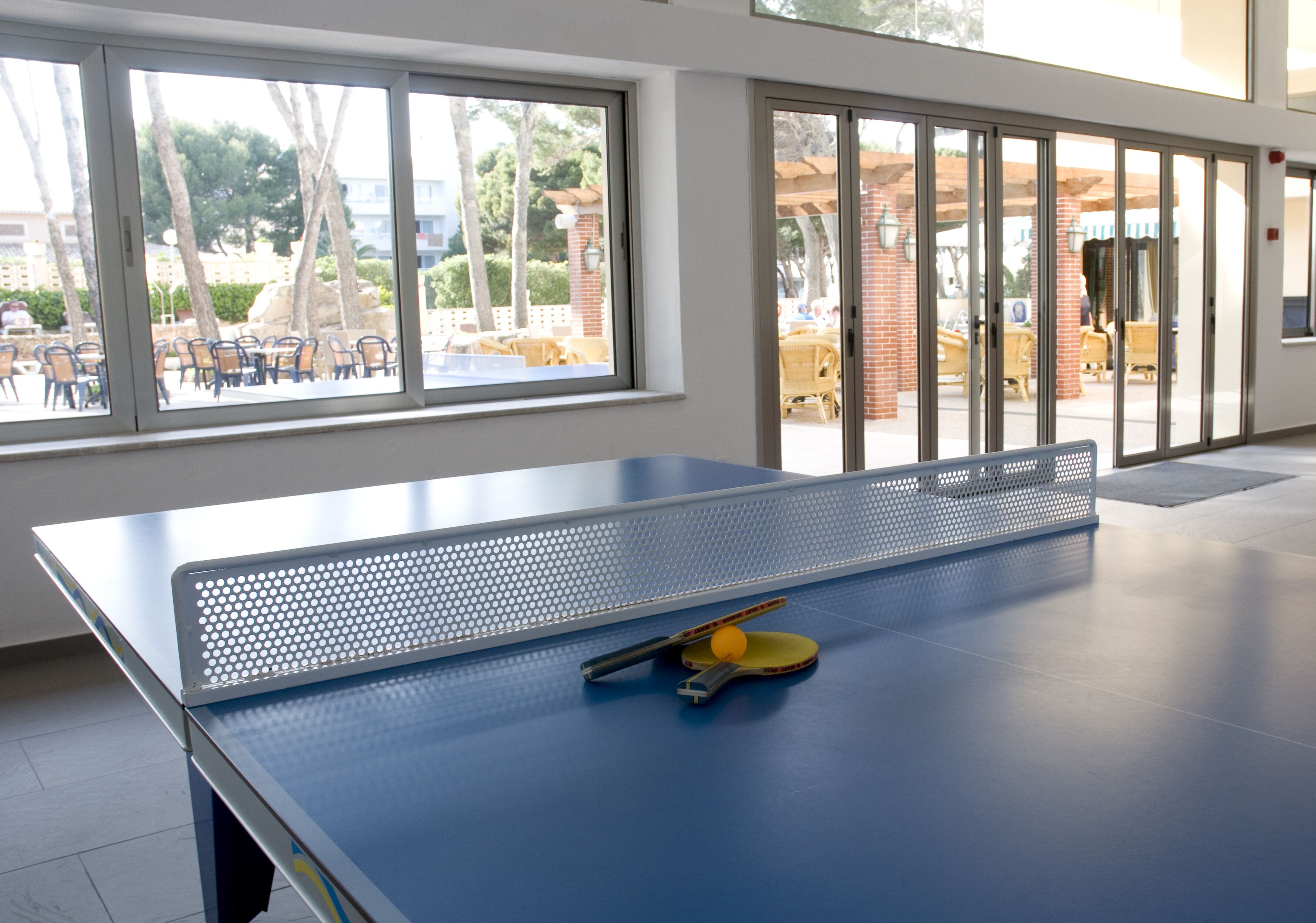 Mesa de ping pong con red, paleta y pelota en una habitación con ventanas amplias y vistas a una terraza.