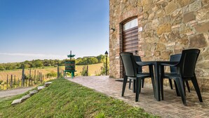Un edificio de piedra con una entrada rústica, un patio empedrado con mesas al aire libre y vistas a un viñedo al fondo.