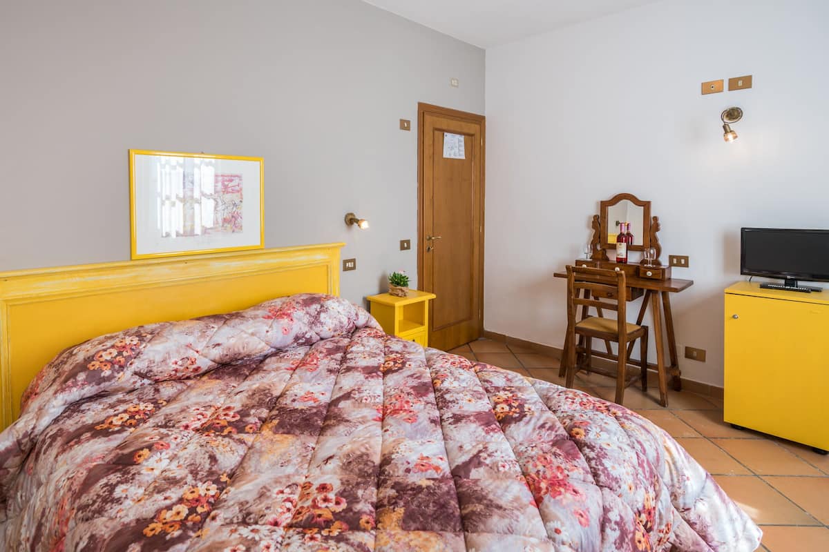 A bedroom with a floral bedspread, a wooden chair, a desk with a mirror, a television, and a yellow cabinet.