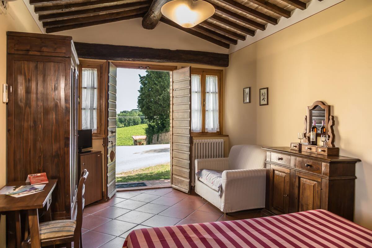 A bedroom with a wooden wardrobe, a bed with a striped bedspread, a chair, a dresser, and a window with a view of greenery outside.