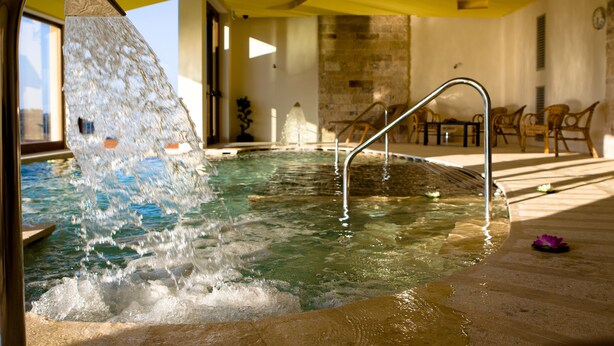 Una bañera de hidromasaje con agua cayendo sobre un borde metálico, rodeada de baldosas de piedra y una terraza de madera.