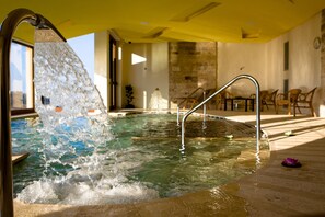 Una bañera de hidromasaje con agua cayendo sobre un borde metálico, rodeada de baldosas de piedra y una terraza de madera.