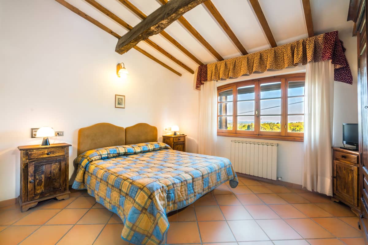 A bedroom with a bed, wooden headboard, bedside tables, a window with curtains, and a radiator.
