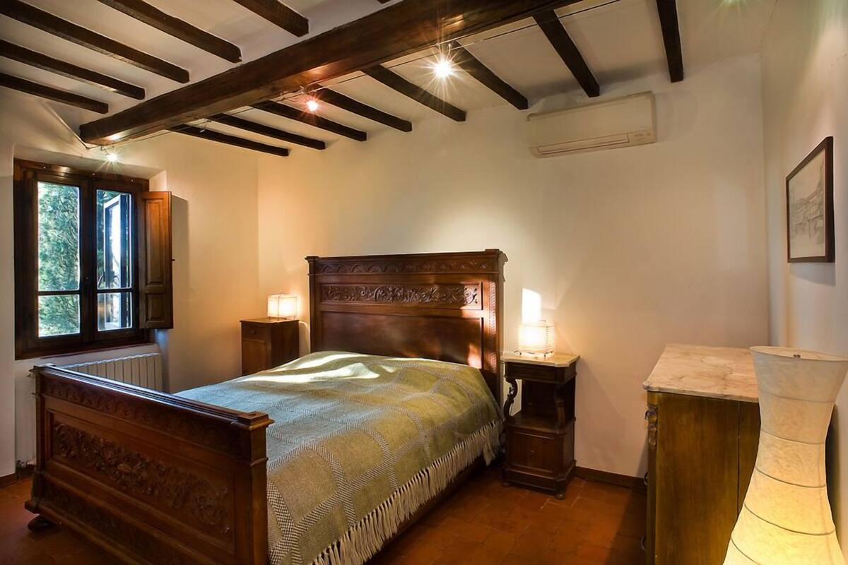 A bedroom with a wooden bed, two bedside tables with lamps, a marble nightstand, and a window with a view of trees.