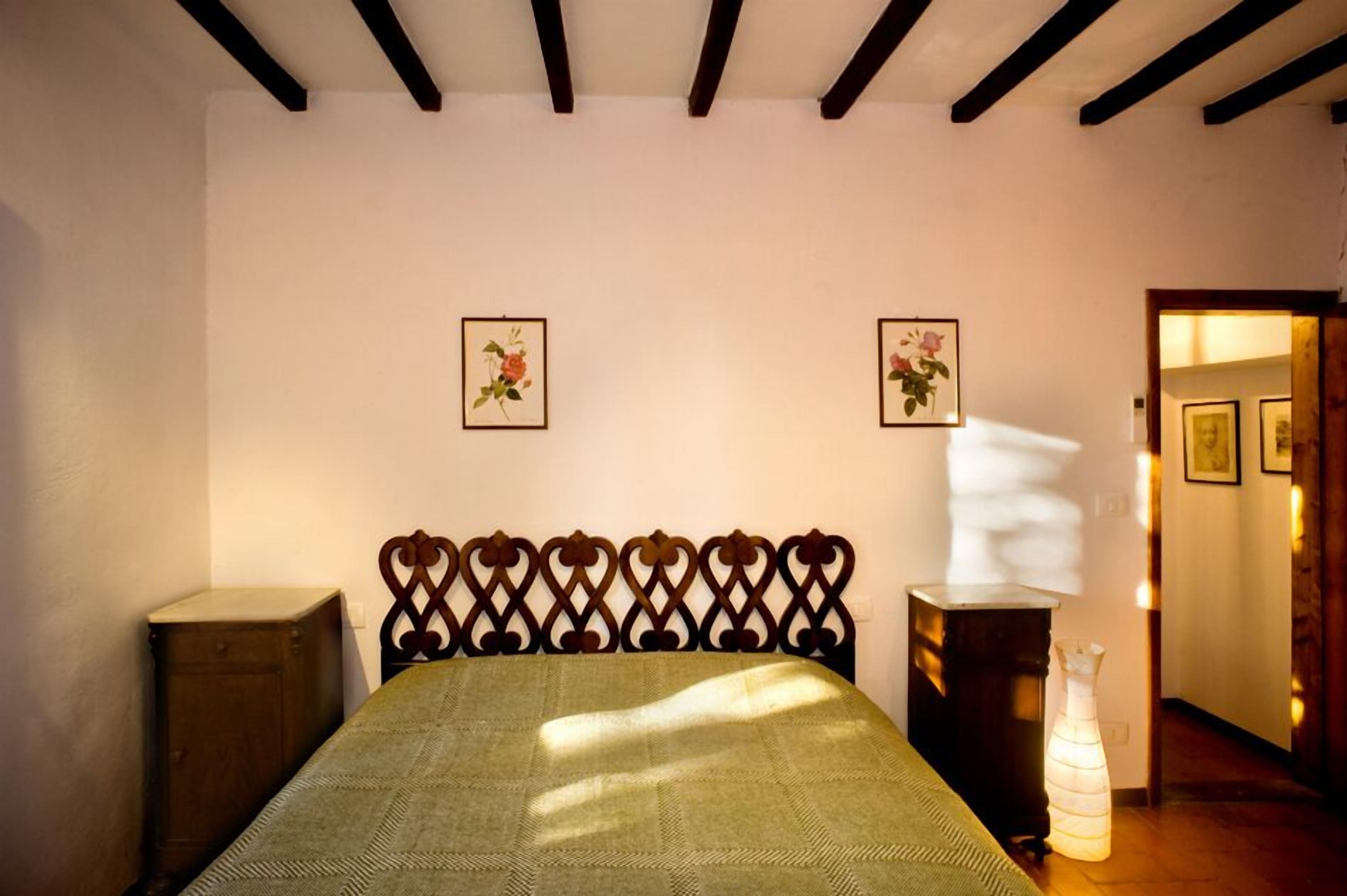 A bedroom with a wooden headboard, a bed with a green bedspread, two framed floral pictures on the wall, a bedside table, and a nightstand with a lamp.