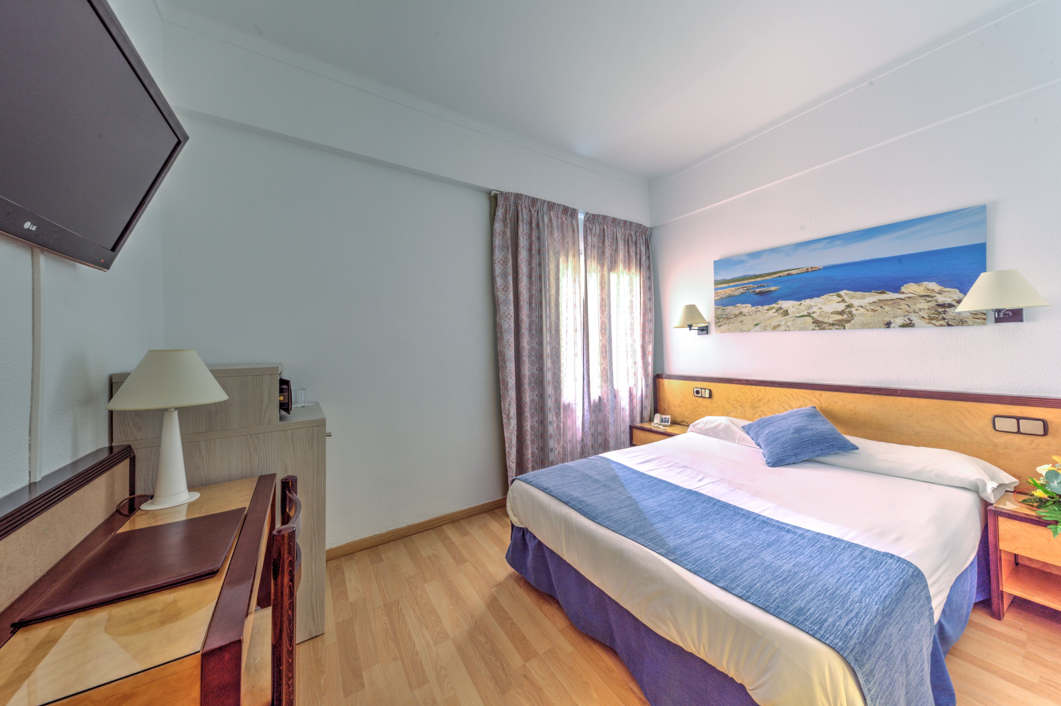 Ein Hotelzimmer mit einem Bett, einem Schreibtisch, einem Fernseher, einem Strandbild und einer Lampe.