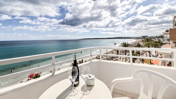 A balcony with a table set for two, overlooking a beach with people swimming and sunbathing, and a cityscape in the distance. - Hotel Voramar Mallorca (Son Servera)