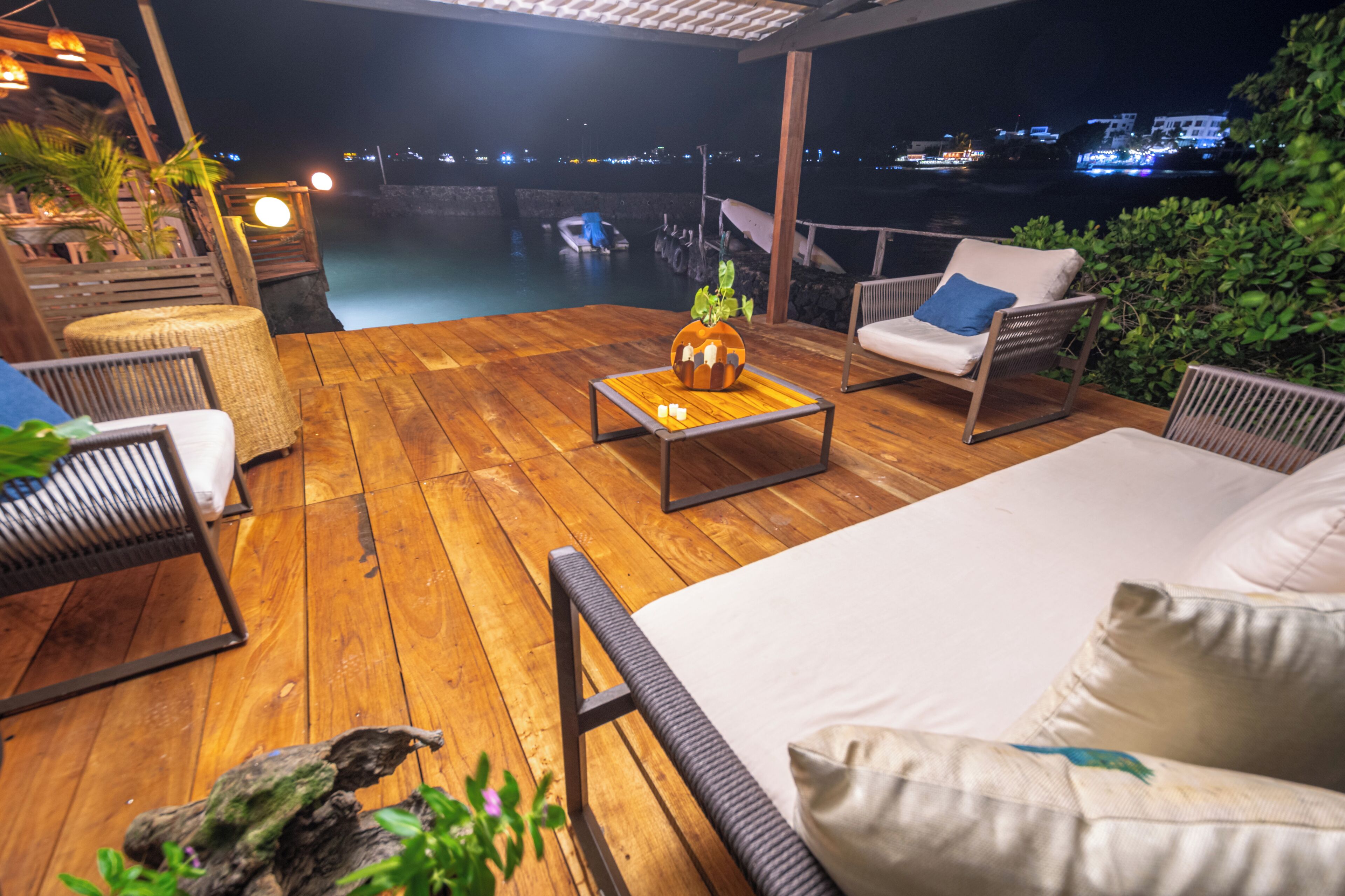 A wooden deck with outdoor furniture, a small table with a decorative item, and a view of the water and city lights in the background.
