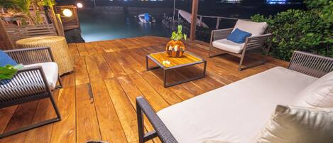 A wooden deck with outdoor furniture, a small table with a decorative item, and a view of the water and city lights in the background.