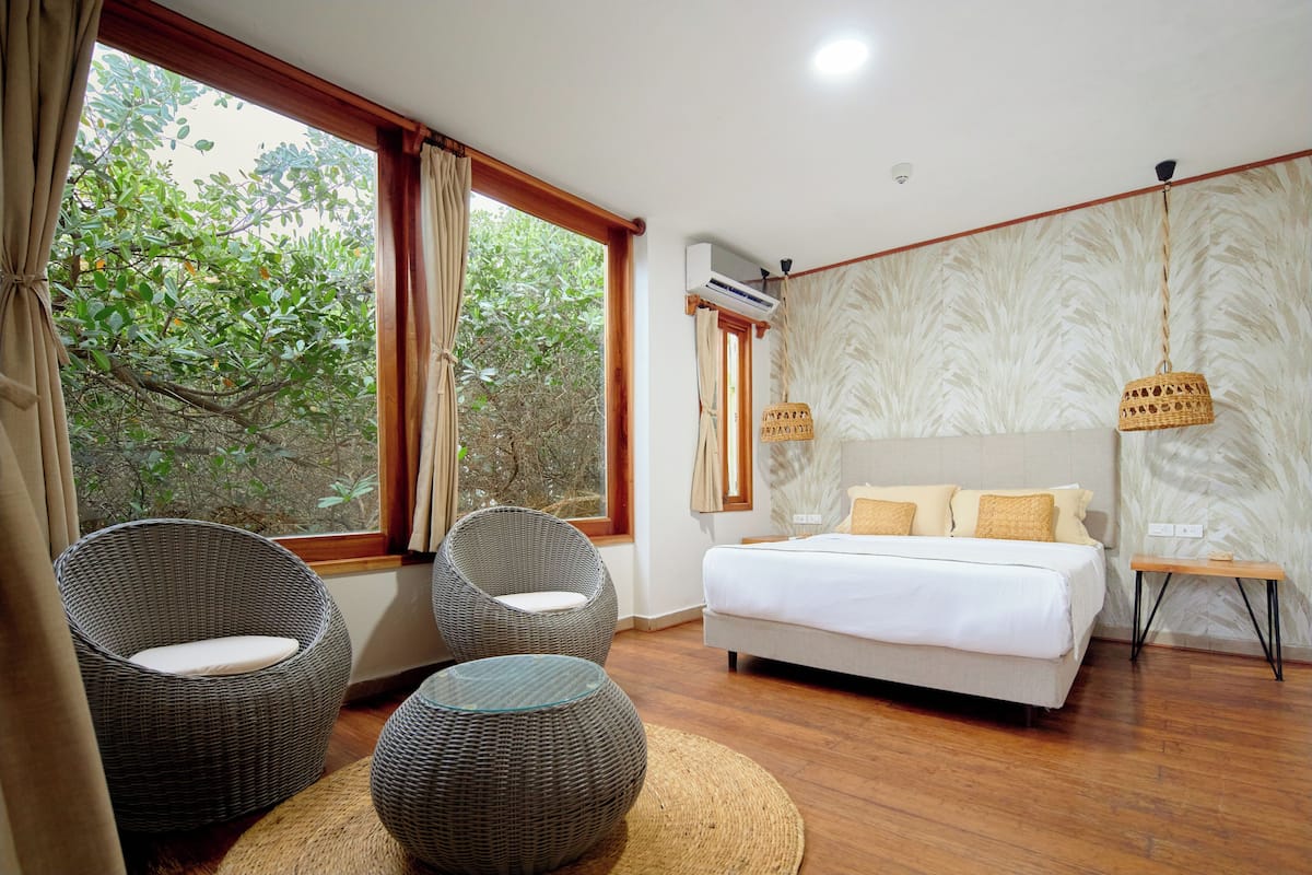 A bedroom with a bed, two wicker chairs, a small table, and a view of trees through large windows.